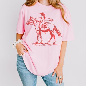以下が含まれることがあります： サーフボードを持った女性が馬に乗っている赤いグラフィックがプリントされた、ピンクのオーバーサイズTシャツです。
