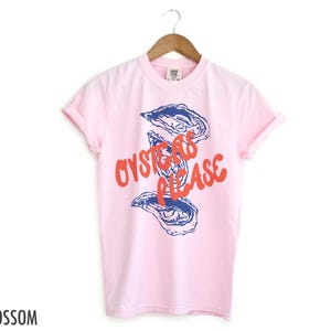 May include: Pink t-shirt with a blue and white illustration of oysters and the text "Oysters Please" in red.