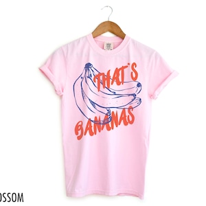 Pu&ograve; includere: Una maglietta rosa con una grafica blu e rossa di banane e il testo "That's Bananas".
