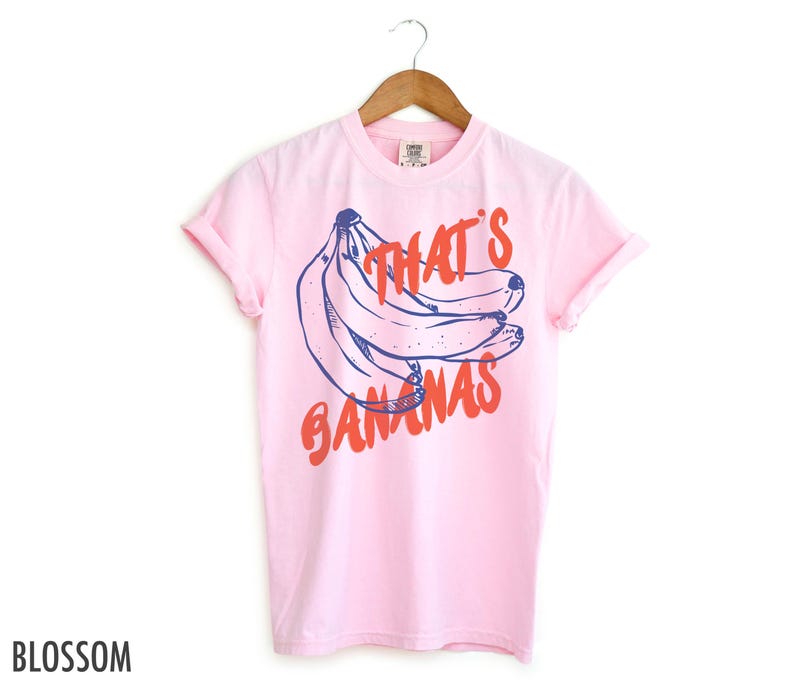 Pu&ograve; includere: Una maglietta rosa con una grafica blu e rossa di banane e il testo "That's Bananas".
