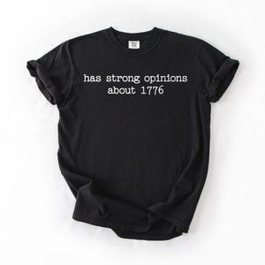 Puede incluir: Camiseta negra con el texto blanco "has strong opinions about 1776". La camiseta tiene cuello redondo, mangas cortas y un corte relajado. La tela parece suave. Es una prenda de vestir informal.