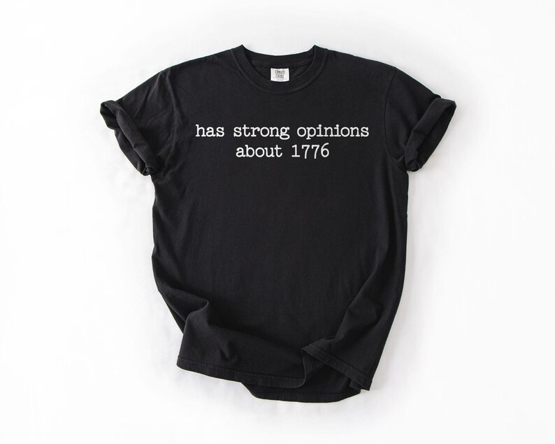 Puede incluir: Camiseta negra con el texto blanco "has strong opinions about 1776". La camiseta tiene cuello redondo, mangas cortas y un corte relajado. La tela parece suave. Es una prenda de vestir informal.