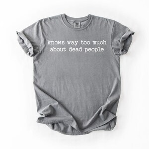 Puede incluir: Camiseta gris con la frase blanca "knows way too much about dead people". La camiseta est&aacute; doblada y tiene las mangas enrolladas. La camiseta est&aacute; hecha de un material suave.
