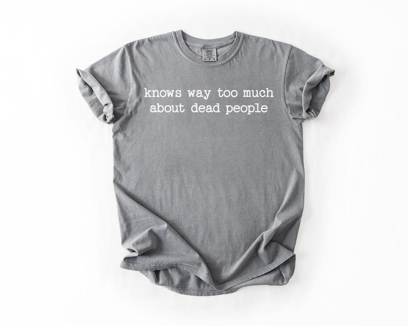 Puede incluir: Camiseta gris con la frase blanca "knows way too much about dead people". La camiseta est&aacute; doblada y tiene las mangas enrolladas. La camiseta est&aacute; hecha de un material suave.