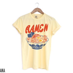 May include: A yellow short-sleeved t-shirt with a graphic of a bowl of ramen noodles and the word "RAMEN" in red letters.