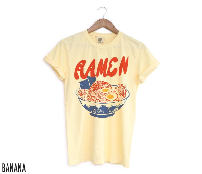 May include: A yellow short-sleeved t-shirt with a graphic of a bowl of ramen noodles and the word "RAMEN" in red letters.