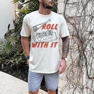 May include: A cream-coloured t-shirt with a graphic of sushi rolls and chopsticks. The text "Roll with it" is printed in red on the shirt.