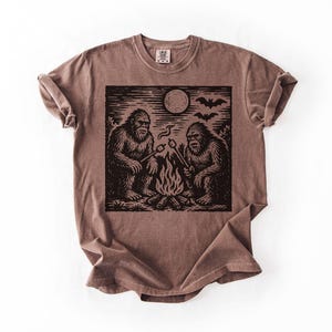 May include: Brown t-shirt featuring a black and white graphic of two Bigfoot creatures roasting marshmallows over a campfire under a full moon. The design includes bats flying in the sky. The shirt has a crew neck and short sleeves.