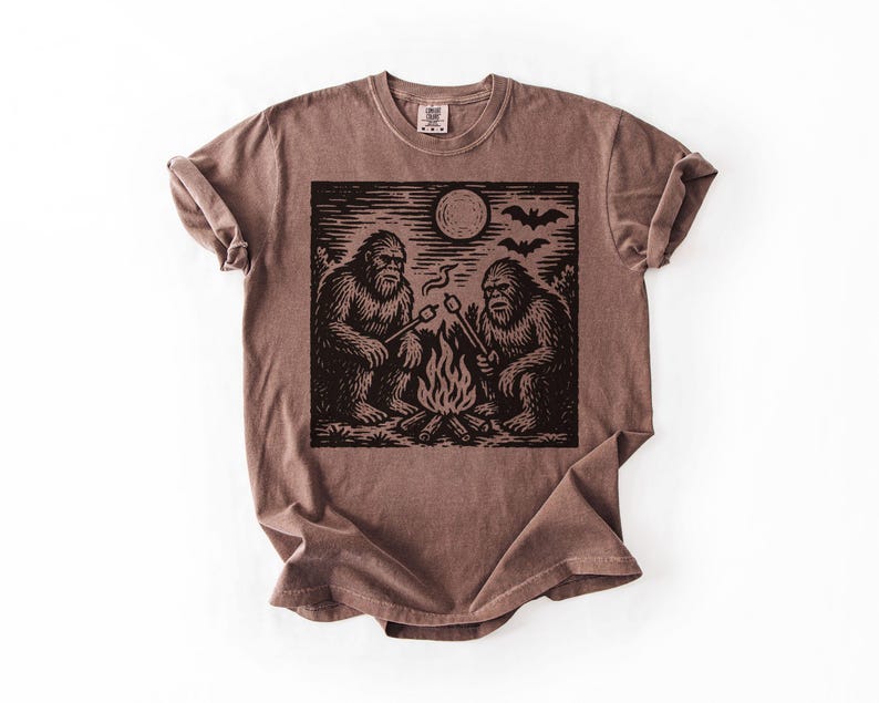 May include: Brown t-shirt featuring a black and white graphic of two Bigfoot creatures roasting marshmallows over a campfire under a full moon. The design includes bats flying in the sky. The shirt has a crew neck and short sleeves.