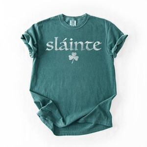 May include: A teal-colored t-shirt with the word "sl&aacute;inte" in white, stylized lettering. Below the word is a white shamrock. The shirt is folded on a white background.