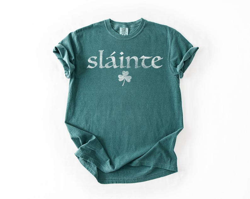 May include: A teal-colored t-shirt with the word "sl&aacute;inte" in white, stylized lettering. Below the word is a white shamrock. The shirt is folded on a white background.