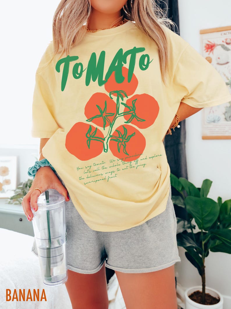 May include: A yellow t-shirt with a green and red tomato graphic and the text "TOMATO" printed on the front. The text below the graphic reads "You say tomato. We say "to-may-to". Let's try all the whole thing off and explore the delicious ways to eat the juicy sun-ripened fruit."