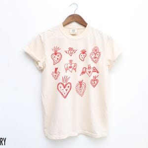 K&ouml;nnte beinhalten: Ein wei&szlig;es T-Shirt mit einem roten Druck verschiedener stilisierter Herzen. Die Herzen haben unterschiedliche Designs, darunter Fl&uuml;gel, Blumen und andere Details.
