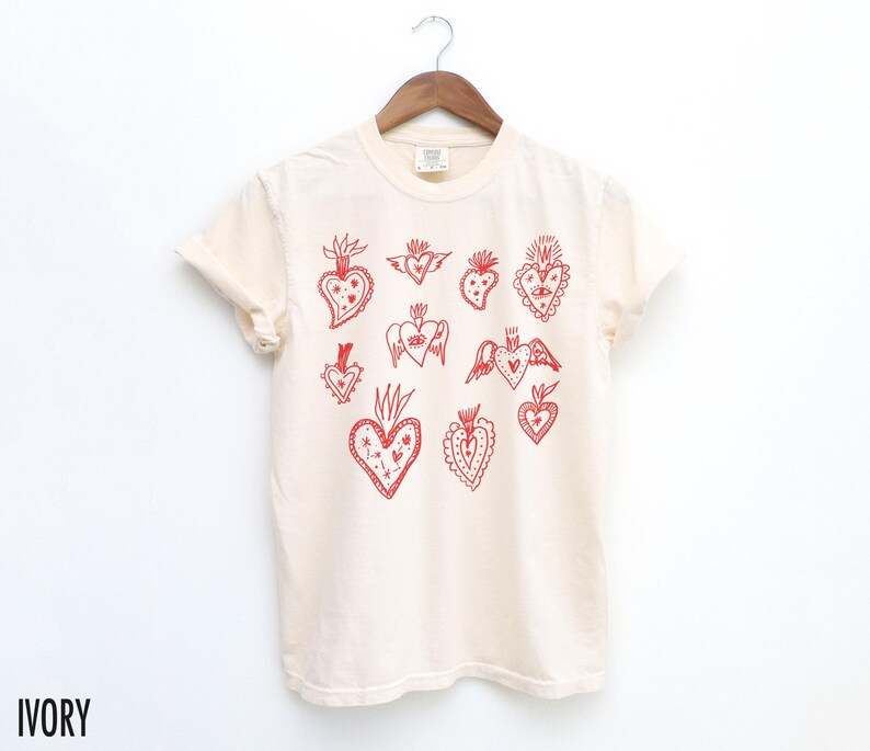 K&ouml;nnte beinhalten: Ein wei&szlig;es T-Shirt mit einem roten Druck verschiedener stilisierter Herzen. Die Herzen haben unterschiedliche Designs, darunter Fl&uuml;gel, Blumen und andere Details.