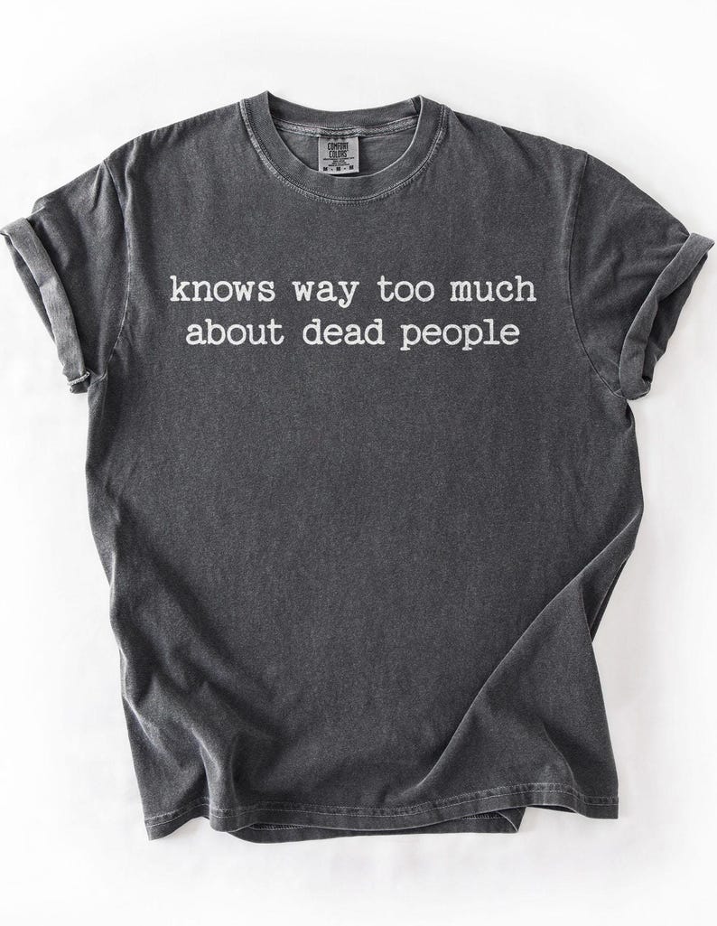 Puede incluir: Camiseta gris oscuro con el texto blanco "knows way too much about dead people". La camiseta tiene cuello redondo y mangas enrolladas. La tela tiene un aspecto vintage ligeramente descolorido.