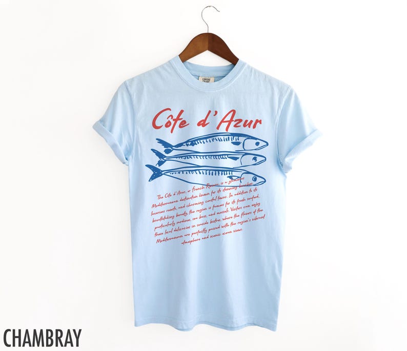 May include: Light blue t-shirt with the text "C&ocirc;te d'Azur" in red script above a blue illustration of three fish. Below the fish is a paragraph of text in red script. The word "CHAMBRAY" is printed in black at the bottom.