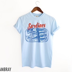K&ouml;nnte beinhalten: Hellblaues T-Shirt mit einem Aufdruck einer Sardinenb&uuml;chse. Der Text "Sardines" ist in Rot &uuml;ber der Dose gedruckt und der Text "We Sardine" ist auf der Dose gedruckt.