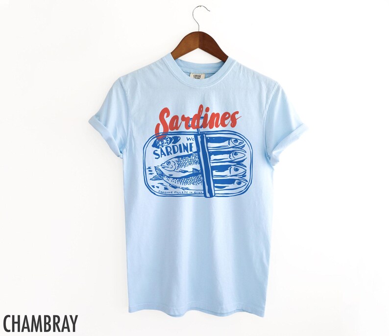 K&ouml;nnte beinhalten: Hellblaues T-Shirt mit einem Aufdruck einer Sardinenb&uuml;chse. Der Text "Sardines" ist in Rot &uuml;ber der Dose gedruckt und der Text "We Sardine" ist auf der Dose gedruckt.