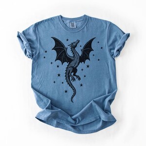 Op de afbeelding: Blauw t-shirt met een zwart drakenontwerp met uitgespreide vleugels en sterren. De draak is gedetailleerd met schubben en een lange staart. Het shirt heeft een ronde hals en korte mouwen.