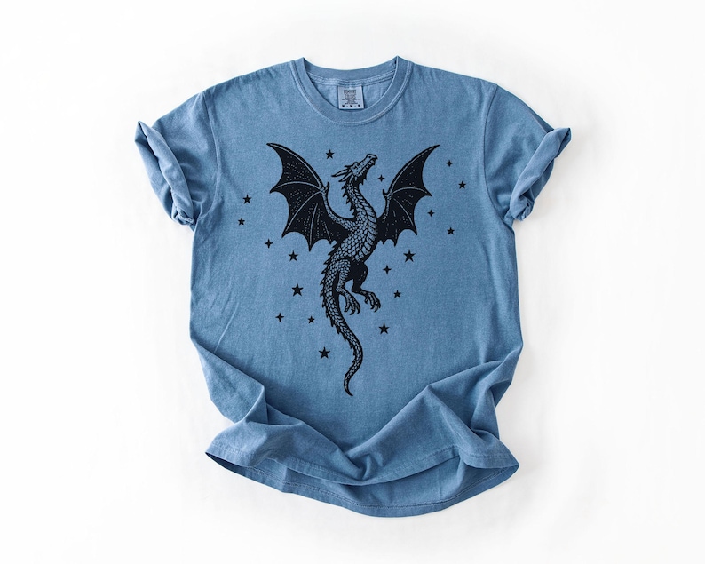Op de afbeelding: Blauw t-shirt met een zwart drakenontwerp met uitgespreide vleugels en sterren. De draak is gedetailleerd met schubben en een lange staart. Het shirt heeft een ronde hals en korte mouwen.