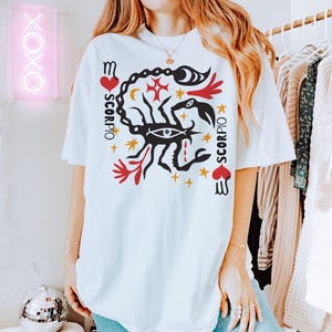 K&ouml;nnte beinhalten: Wei&szlig;es T-Shirt mit einem Grafikdruck eines Skorpions mit einem roten Herzen und dem Wort "Scorpio", das vertikal auf beiden Seiten des Skorpions geschrieben ist.