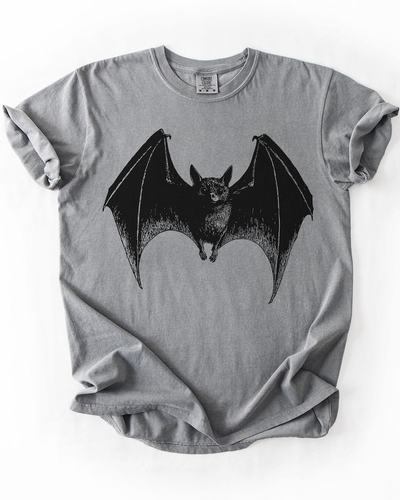 May include: A gray t-shirt featuring a detailed black illustration of a bat with outstretched wings. The bat is centered on the shirt, showcasing its wingspan. The shirt has a classic crew neck and short sleeves.