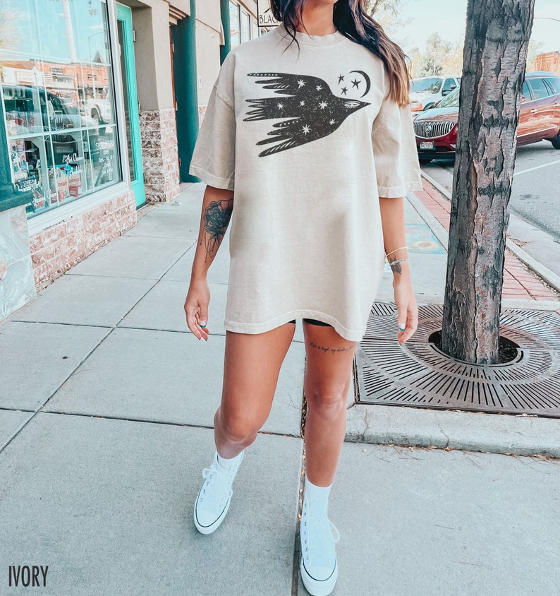May include: Ivory-colored oversized t-shirt featuring a black graphic of a bird with stars and a crescent moon. The shirt has short sleeves and a relaxed fit. The word "IVORY" is printed at the bottom of the image.