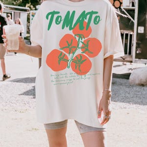 May include: A white oversized t-shirt with a tomato graphic and the text "TOMATO" in green. The text below the graphic reads "You say tomato, we say..." and continues with a description of the tomato. The shirt is labeled "IVORY".