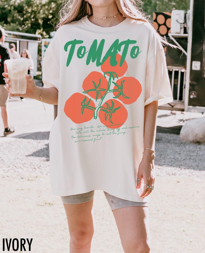 May include: A white oversized t-shirt with a tomato graphic and the text "TOMATO" in green. The text below the graphic reads "You say tomato, we say..." and continues with a description of the tomato. The shirt is labeled "IVORY".