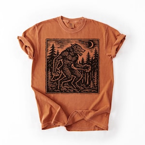 Puede incluir: Camiseta de color naranja quemado con un gr&aacute;fico negro de un hombre lobo en un bosque bajo una luna creciente. El dise&ntilde;o est&aacute; enmarcado dentro de un borde rectangular. La camiseta tiene cuello redondo y mangas enrolladas.
