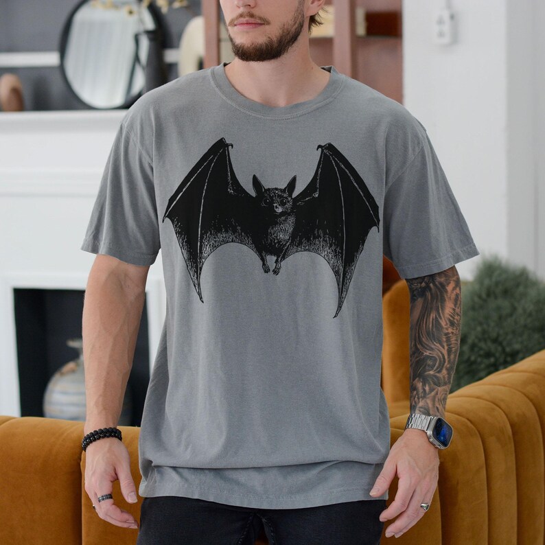 May include: Gray t-shirt featuring a large black bat graphic. The bat is depicted with outstretched wings, centered on the shirt. The shirt has short sleeves and a relaxed fit. The overall style is casual and suitable for everyday wear.