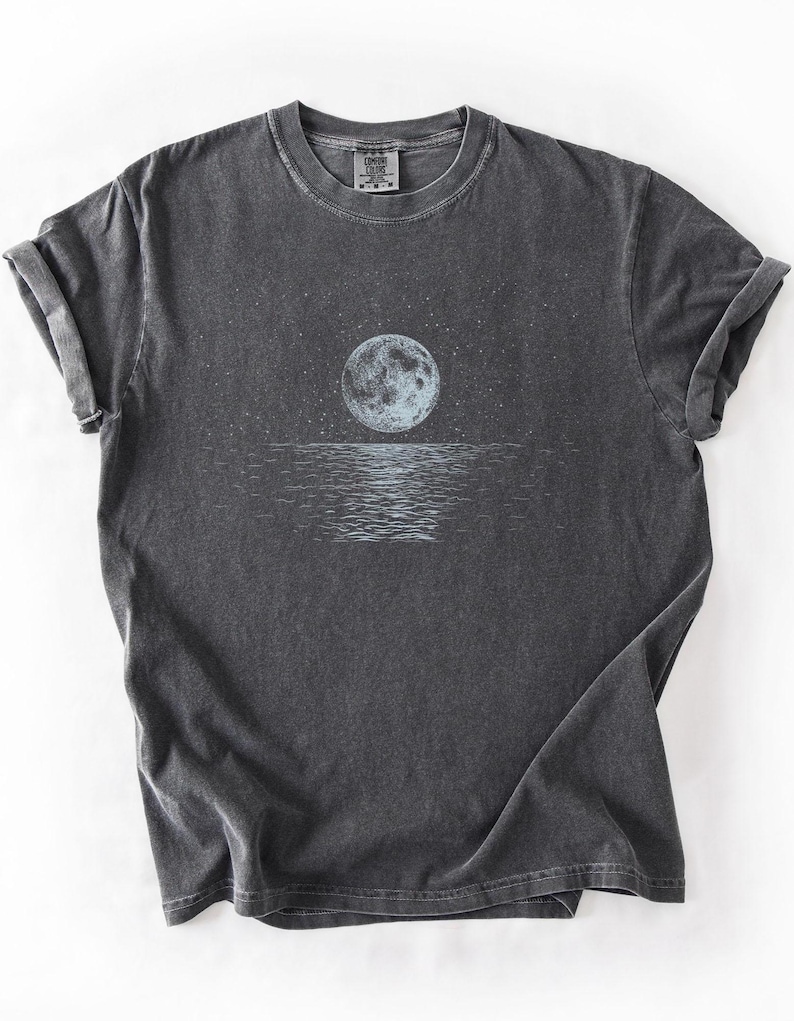 May include: Dark gray t-shirt featuring a full moon over water with stars in the sky. The moon is light blue and the water has a reflective pattern. The shirt has rolled-up sleeves and a crew neck.