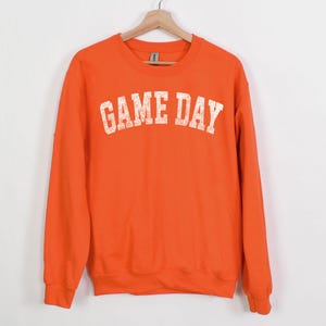 May include: An orange sweatshirt with the text "GAME DAY" in a distressed white font. The crew neck sweatshirt is hanging on a wooden hanger against a white background. The word "ORANGE" is printed at the bottom left.