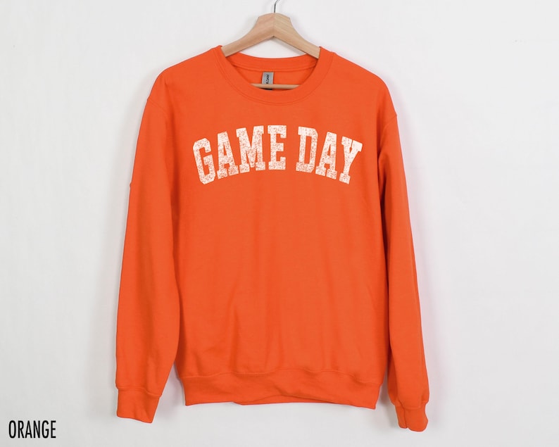 May include: An orange sweatshirt with the text "GAME DAY" in a distressed white font. The crew neck sweatshirt is hanging on a wooden hanger against a white background. The word "ORANGE" is printed at the bottom left.