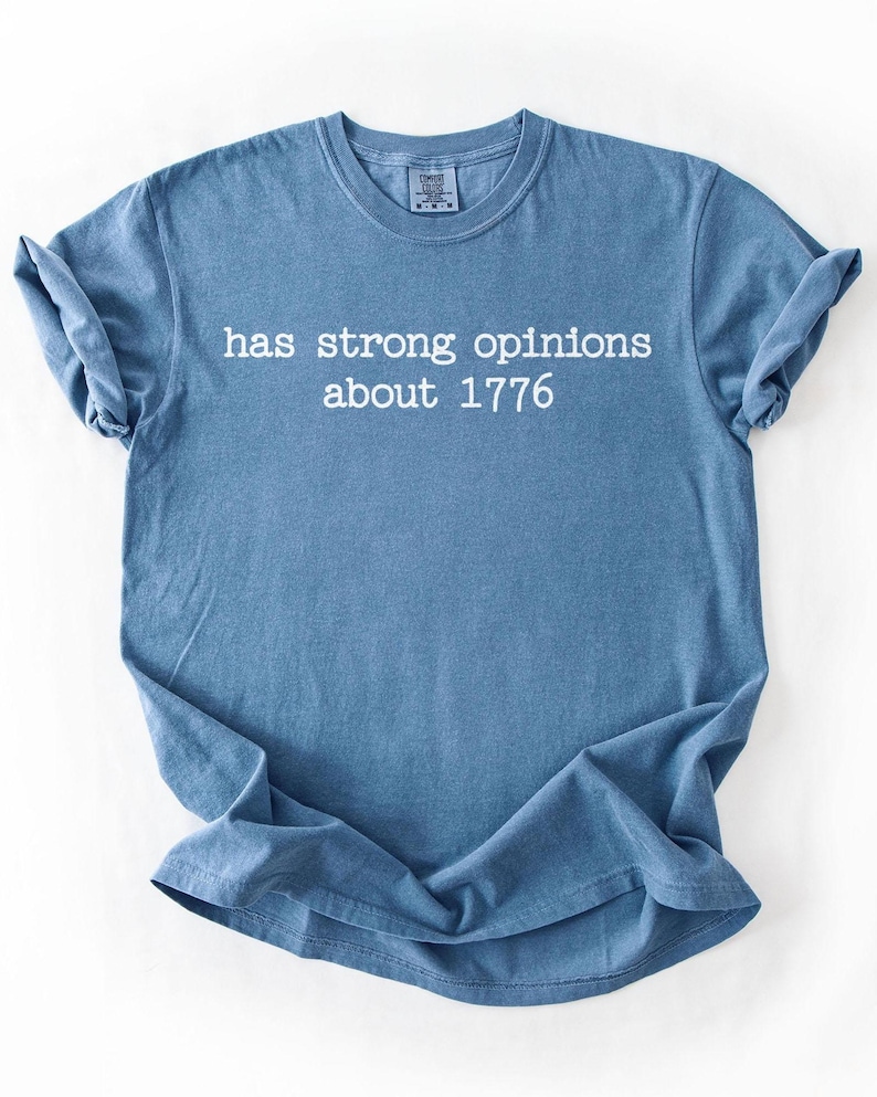 Puede incluir: Camiseta azul con el texto blanco "has strong opinions about 1776". La camiseta tiene cuello redondo y mangas enrolladas. La tela parece suave.