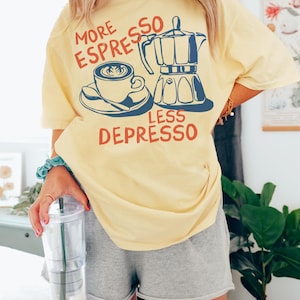 May include: A yellow t-shirt with the text "More Espresso Less Depresso" in red and blue lettering. The design features a coffee cup and a coffee pot.