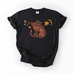 May include: Black t-shirt featuring an orange tiger design with stars and a crescent moon. The tiger has the word "RAA" above it. The shirt is made of a soft material and has short sleeves.