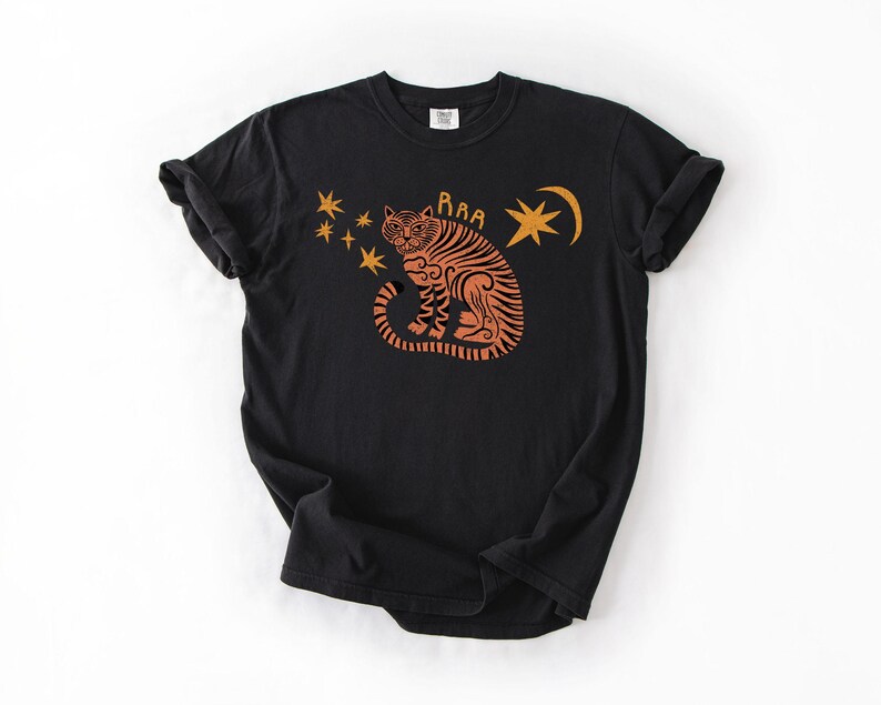 May include: Black t-shirt featuring an orange tiger design with stars and a crescent moon. The tiger has the word "RAA" above it. The shirt is made of a soft material and has short sleeves.