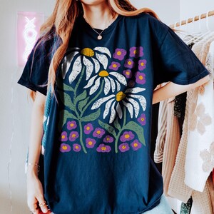 May include: Navy blue t-shirt featuring a floral design with large white daisies with yellow centers, green leaves, and small purple flowers. The shirt has a relaxed fit and short sleeves. The design is a bold, graphic print.