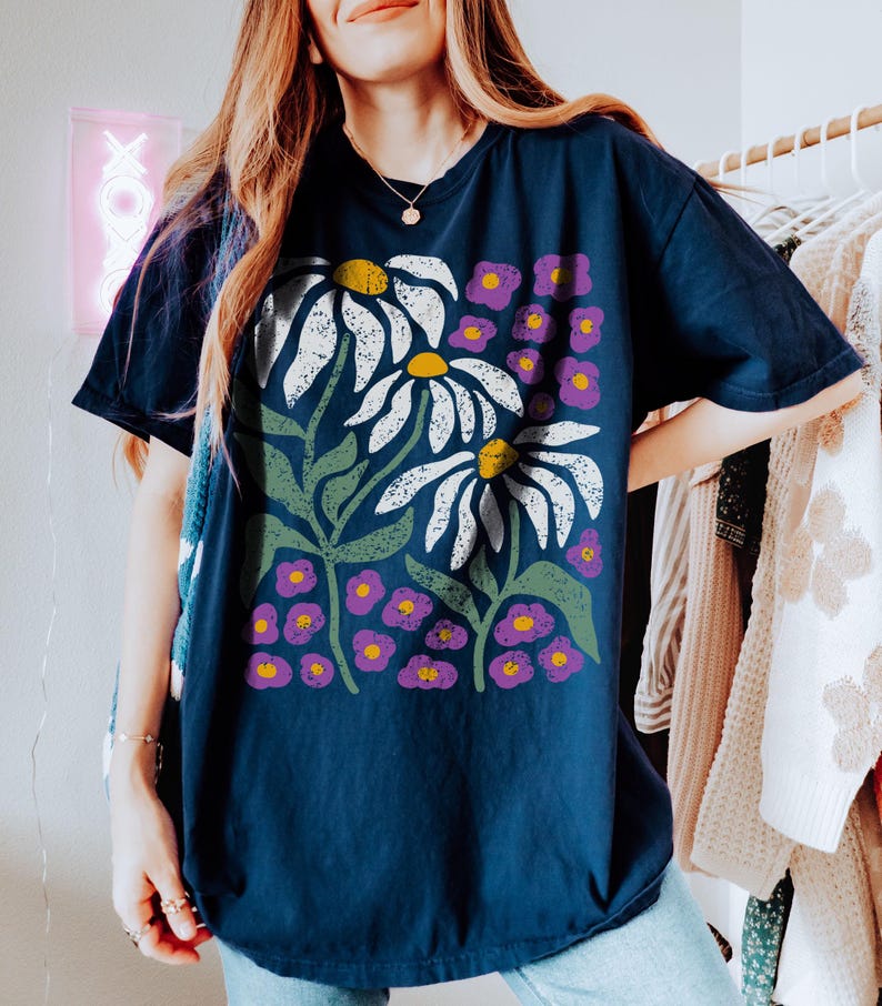 May include: Navy blue t-shirt featuring a floral design with large white daisies with yellow centers, green leaves, and small purple flowers. The shirt has a relaxed fit and short sleeves. The design is a bold, graphic print.