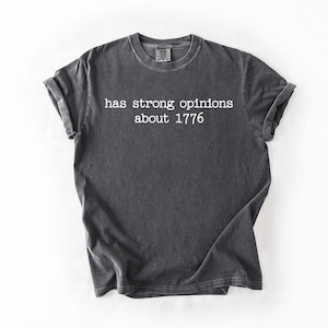 Puede incluir: Camiseta gris oscuro con mangas enrolladas. La camiseta presenta el texto blanco: "has strong opinions about 1776". La camiseta est&aacute; hecha de un material suave y es adecuada para ropa casual.