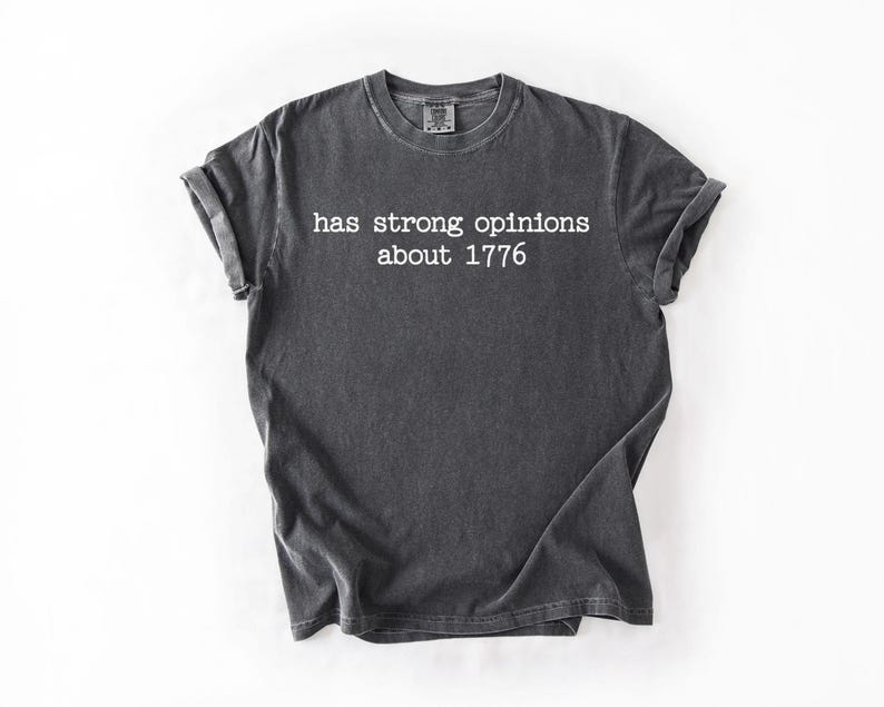 Puede incluir: Camiseta gris oscuro con mangas enrolladas. La camiseta presenta el texto blanco: "has strong opinions about 1776". La camiseta est&aacute; hecha de un material suave y es adecuada para ropa casual.