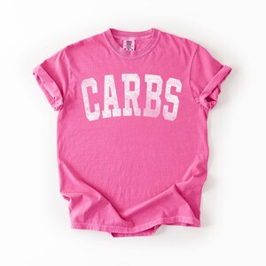 May include: A bright pink t-shirt with the word "CARBS" printed in large, distressed white letters across the front. The shirt has short sleeves and a crew neck. The fabric is slightly wrinkled, suggesting a casual style.