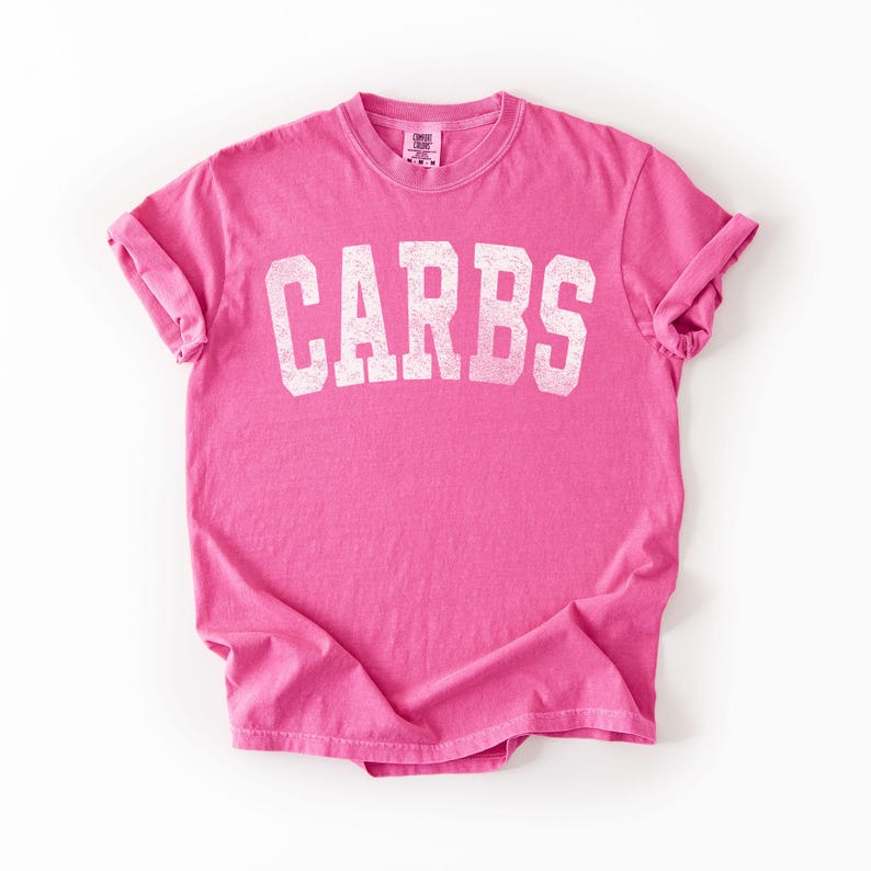 May include: A bright pink t-shirt with the word "CARBS" printed in large, distressed white letters across the front. The shirt has short sleeves and a crew neck. The fabric is slightly wrinkled, suggesting a casual style.