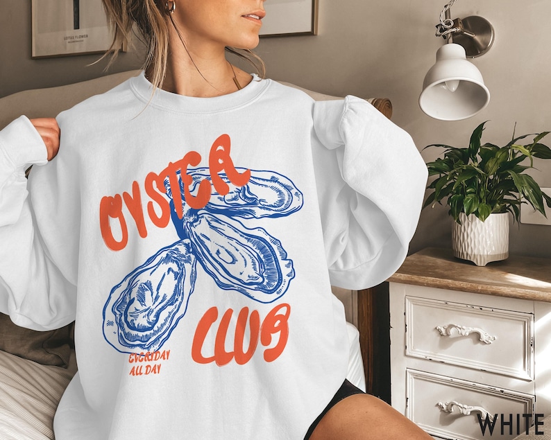 K&ouml;nnte beinhalten: Wei&szlig;er Pullover mit einer blauen und orangenen Grafik von Austern und dem Text "Oyster Club Everyday All Day".