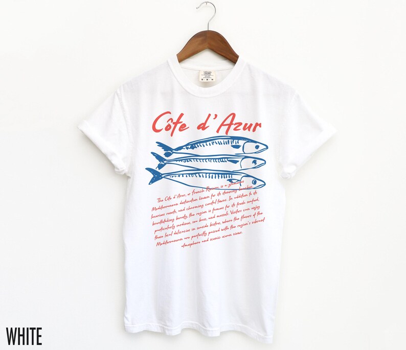 May include: White t-shirt with a graphic design featuring three blue fish and the text "C&ocirc;te d'Azur" in red. Below the text is a paragraph of red text. The shirt is hanging on a wooden hanger.