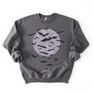 May include: A dark grey sweatshirt with a distressed graphic of a full moon and bats. The bats are black, and the moon is a faded purple. The sweatshirt has long sleeves and a crew neck, perfect for Halloween.