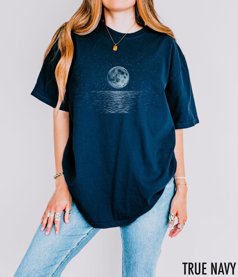 May include: A navy blue t-shirt featuring a detailed illustration of a full moon reflecting on water under a starry sky. The text "TRUE NAVY" is printed in white at the bottom right corner of the image.