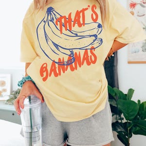 Pu&ograve; includere: Una maglietta gialla con una grafica di banane e il testo "That's Bananas".