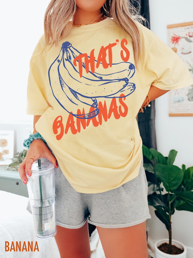 Pu&ograve; includere: Una maglietta gialla con una grafica di banane e il testo "That's Bananas".